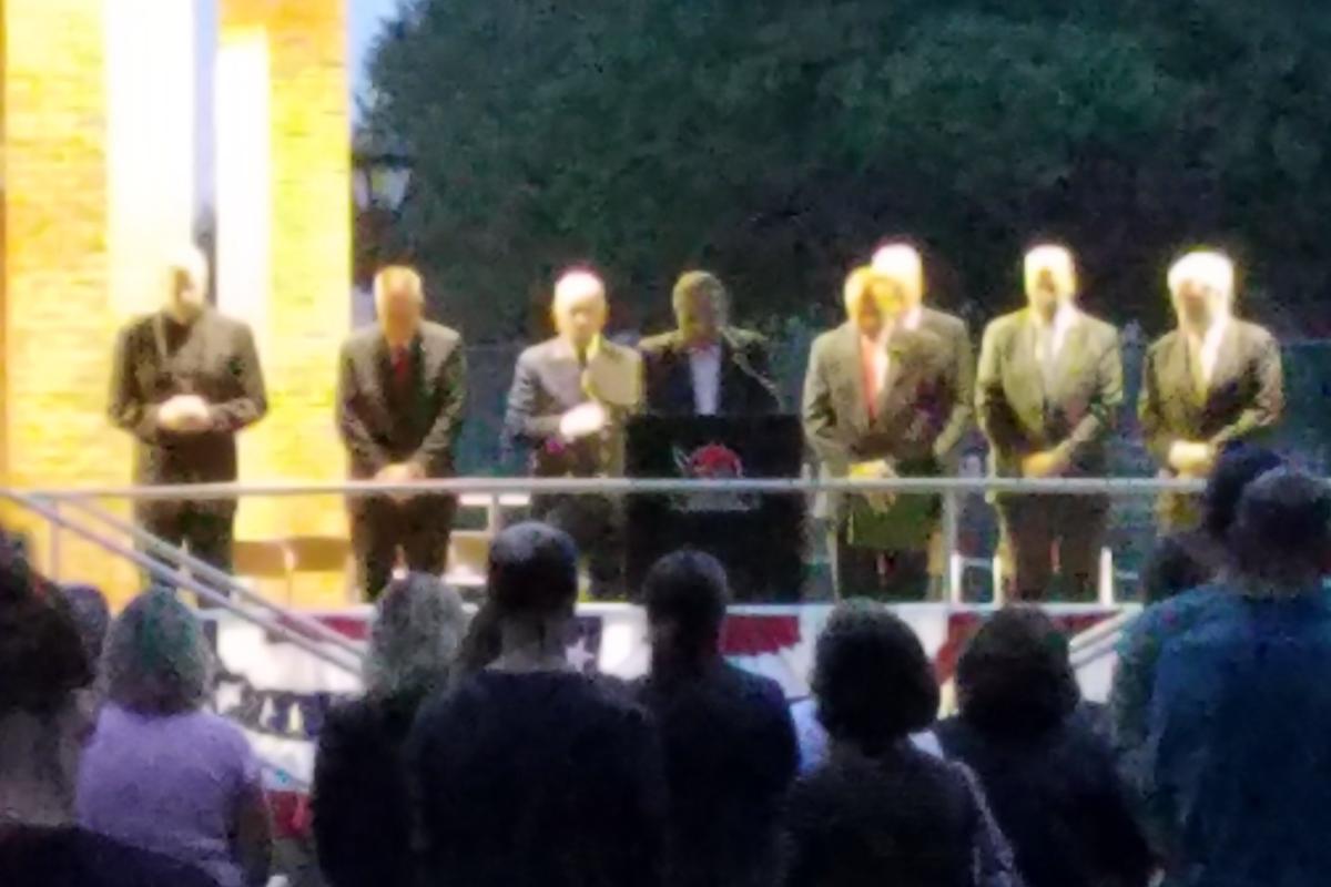 Eight men on stage standing with heads down to say a prayer
