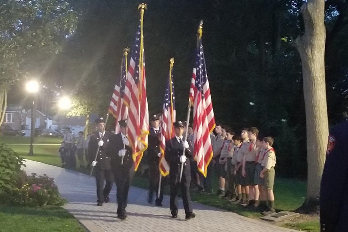 Four uniformed firemen carrying flags walking on path, boy scouts lined up on grass at edge of path