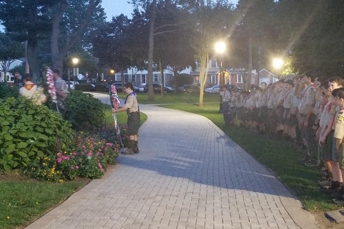 Paved park path, boy scouts lined up along path, one scout lifting red white blue wreath and stand, large trees in background