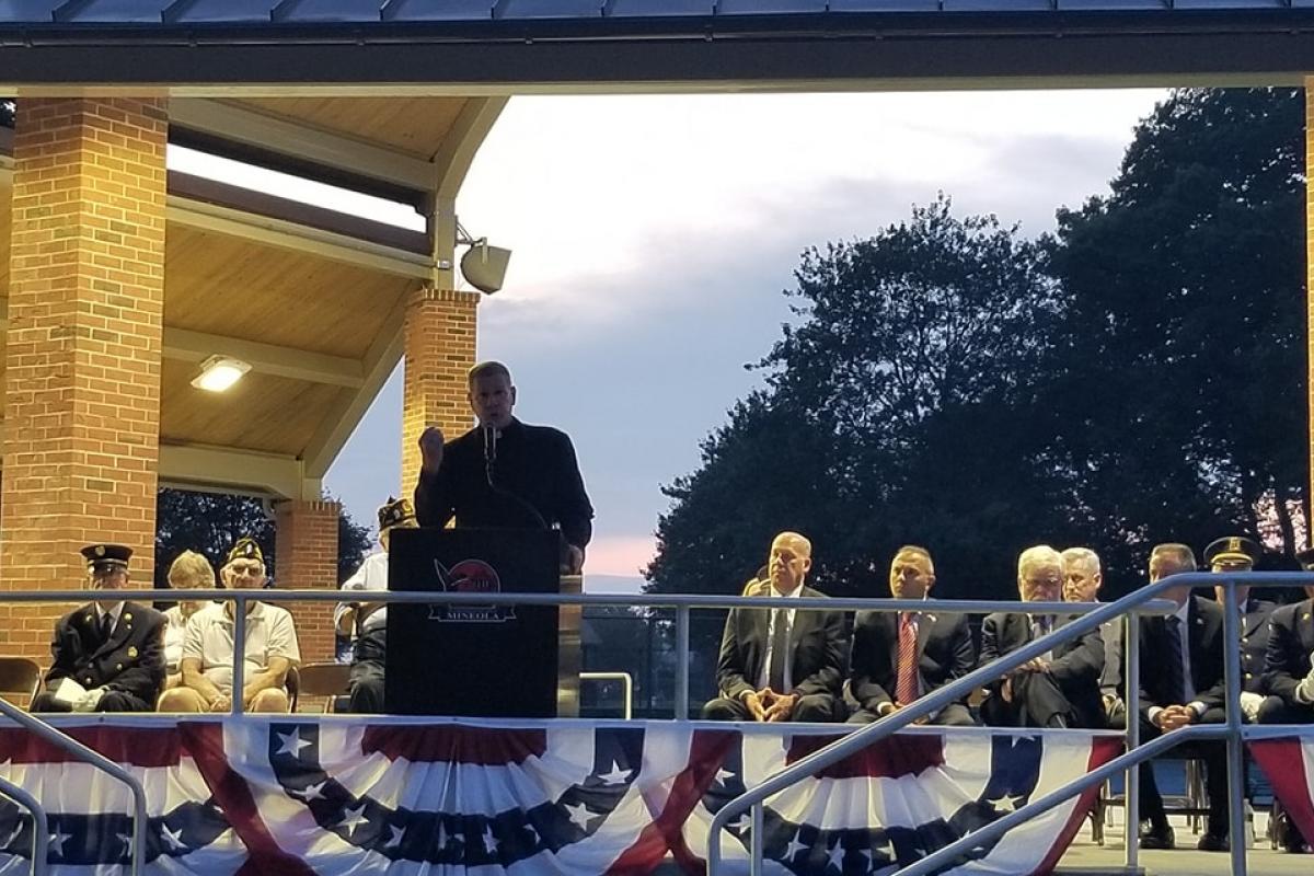 Priest at podium, people seated on either side of podium, red white blue bunting, sun setting, large trees in distance