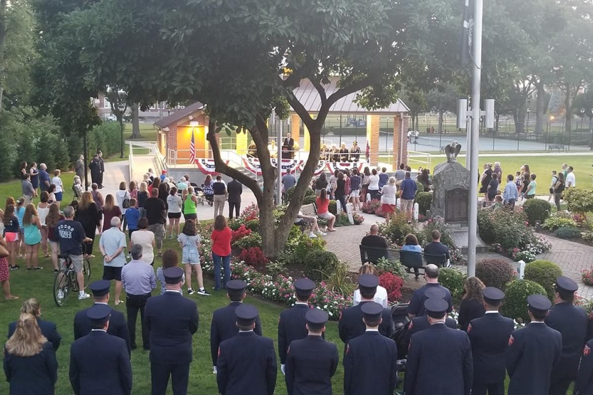 View of event from back, stage in distance, backs of fireman and audience, large tree in center