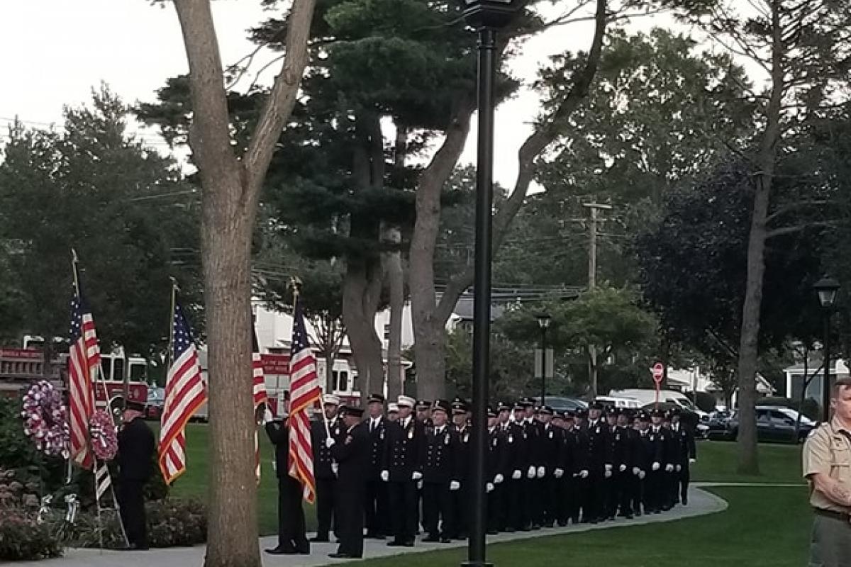 Firemen wearing blue uniforms lined up on park walkway first two holding American flags trees and lampost