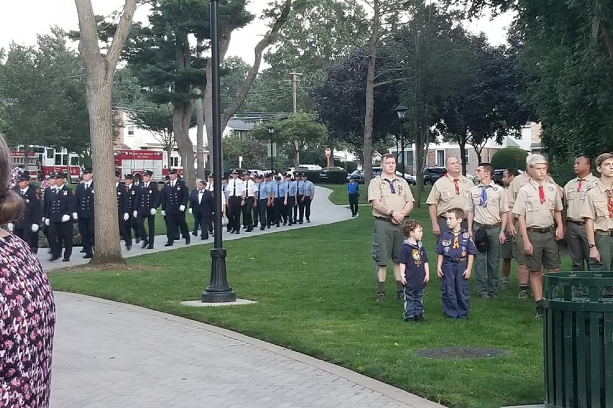 Group of standing boy scouts and cub scouts watching uniformed firemen march into park