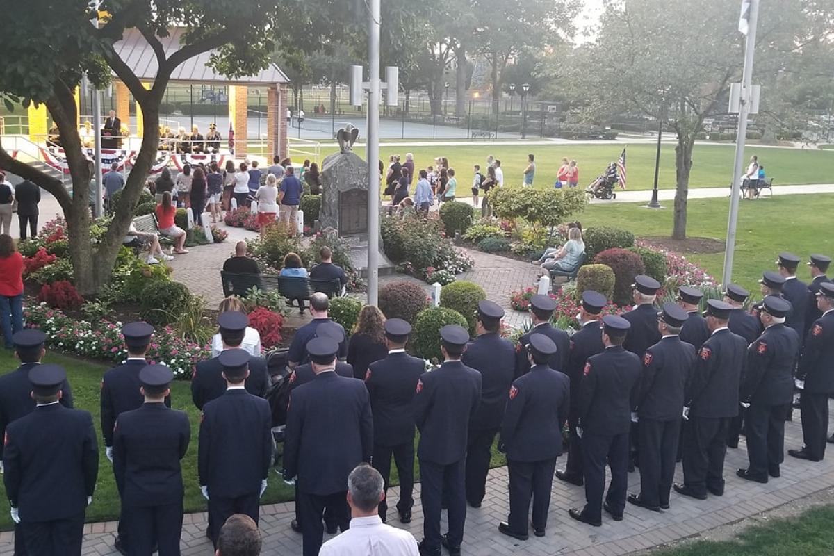 View of event from back, stage in distance, backs of fireman and audience, large tree on left, monument in center