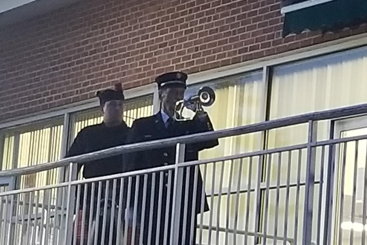 Uniformed elderly man playing bugle with younger man and building behind him