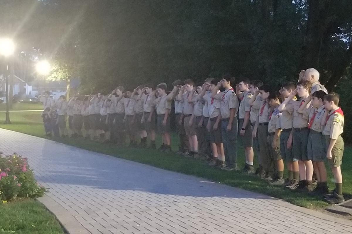 Boy scouts and troop leaders lined up on grass at edge of walking path saluting, trees in background