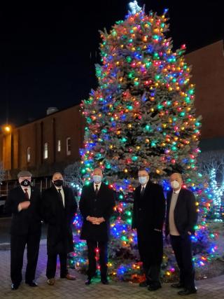 Nighttime, lit Christmas tree with five men in suits wearing masks in front of tree